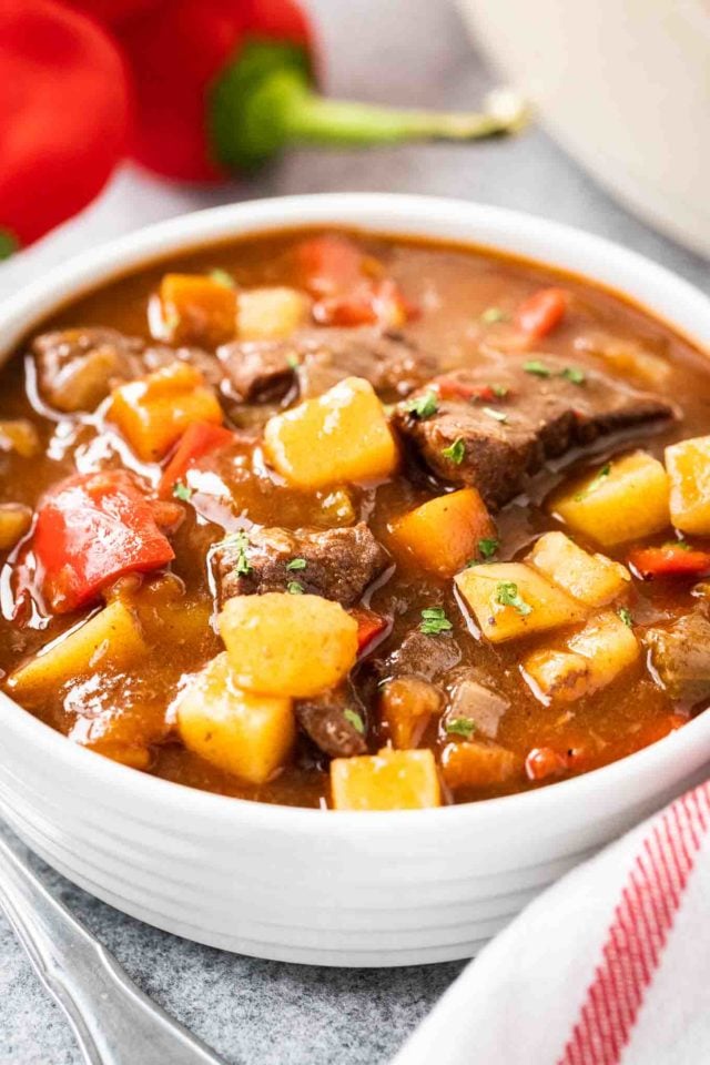 Un bol blanc de goulash, contenant des poivrons rouges, du bœuf et des pommes de terre, garni de persil. Il y a un torchon blanc avec des rayures rouges et quelques poivrons rouges à côté.