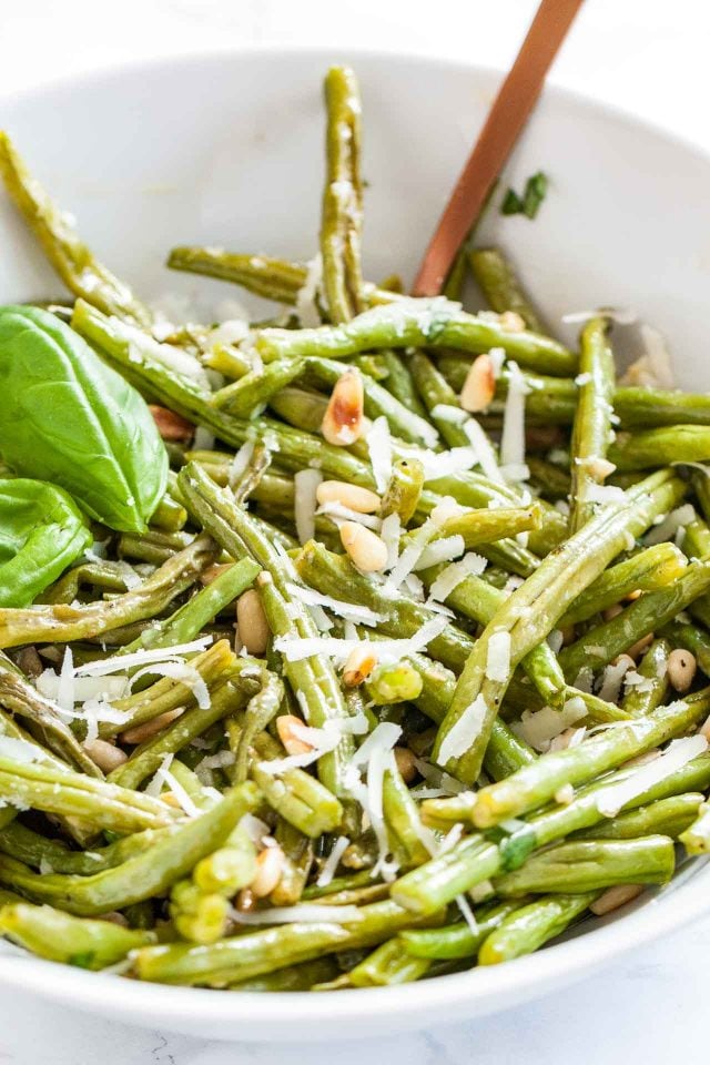 Gros plan d'un bol blanc de haricots verts rôtis au four surmontés de pignons de pin grillés, de Parmigiano et de feuilles de basilic.