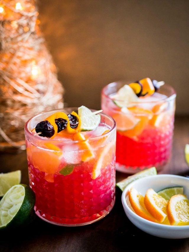 Deux verres de Moscow mule aux oranges et canneberges avec des brochettes de fruits. Il y a un petit bol blanc de quartiers de citron vert et d'orange ainsi que des citrons verts coupés à côté et une décoration de Noël en arrière-plan.