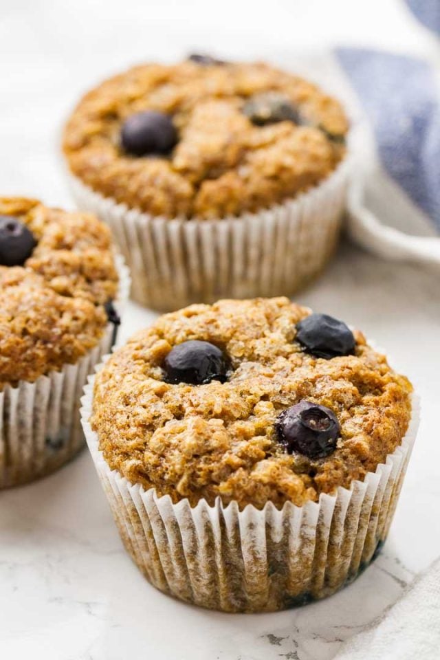 Trois muffins à la banane et aux myrtilles sur une surface en marbre à côté d'un torchon blanc et bleu.