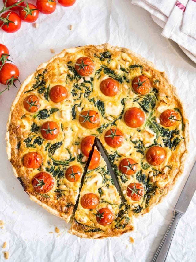 Quiche aux épinards et aux tomates sur un torchon blanc avec un couteau, un torchon rouge et blanc et quelques tomates à côté. Une part a été coupée mais est encore à sa place.