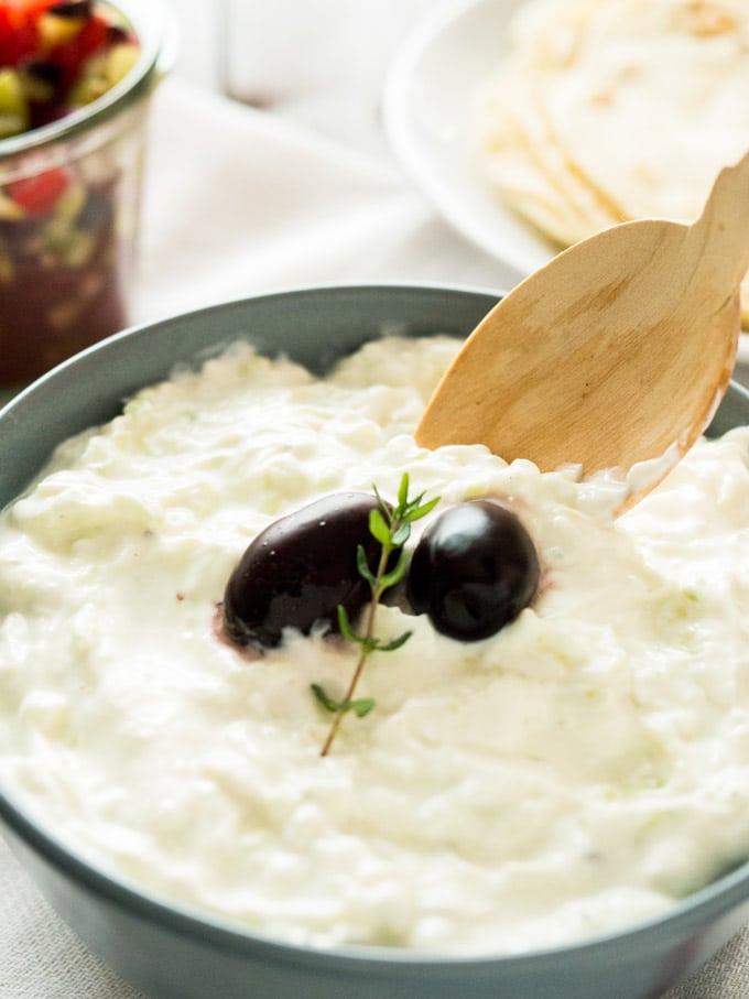 Gros plan d'un bol gris ardoise de Tzatziki, avec deux olives noires et un brin de thym avec une cuillère en bois dedans sur un torchon blanc. Il y a une assiette blanche de tortillas et un bocal de salade grecque en arrière-plan.