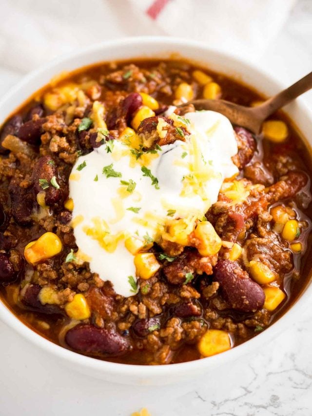 vue de dessus d'un bol blanc de chili con carne, surmonté de crème aigre, de coriandre et de fromage râpé avec une cuillère en bronze.