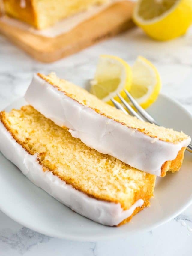 Deux tranches de gâteau au citron moelleux avec un glaçage au citron reposant sur une assiette blanche avec une fourchette garnie de deux quartiers de citron. Une planche à découper en bois avec le reste du gâteau est en arrière-plan.