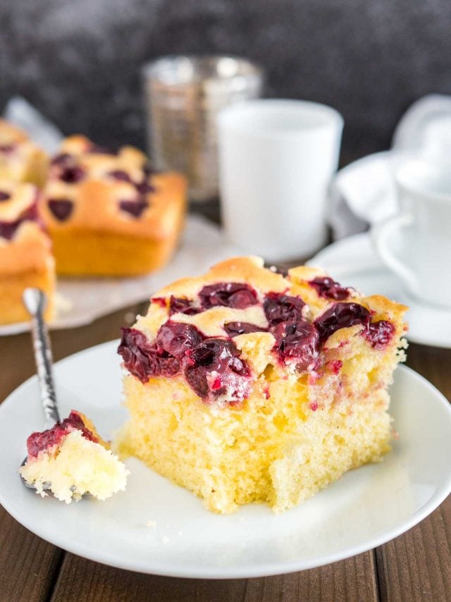 Un morceau de gâteau aux cerises sur une assiette blanche avec une bouchée prise par une cuillère. Le reste du gâteau est en arrière-plan ainsi qu'une tasse blanche.