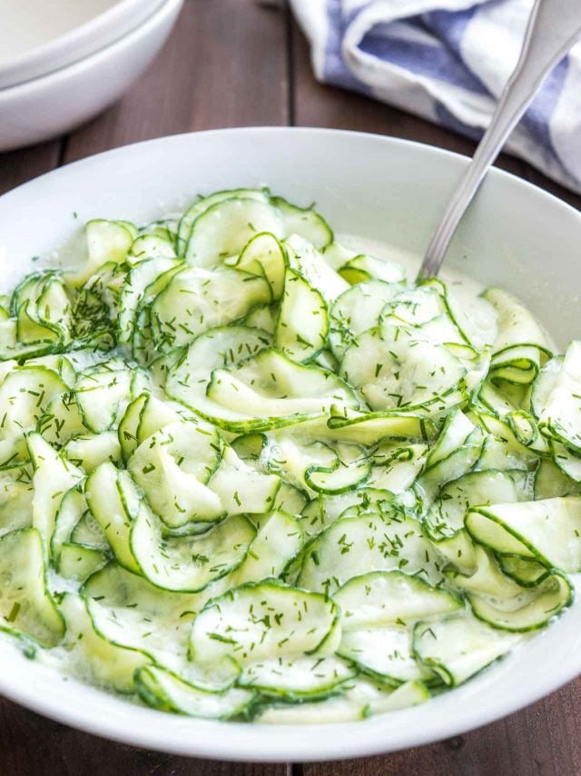 Salade de concombre allemande dans un bol de salade blanc garni d'aneth sur une table en bois avec une cuillère dedans.