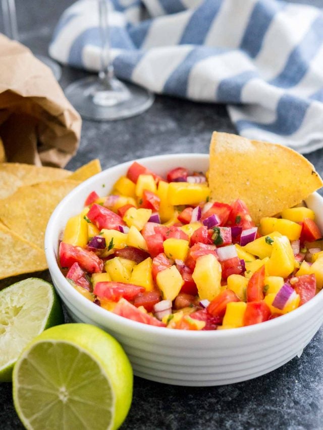 Un bol blanc contenant de la salsa aux tomates et à la mangue avec une chips à l'intérieur sur une surface sombre à côté de deux citrons verts, un sachet de nachos et un torchon blanc et bleu.