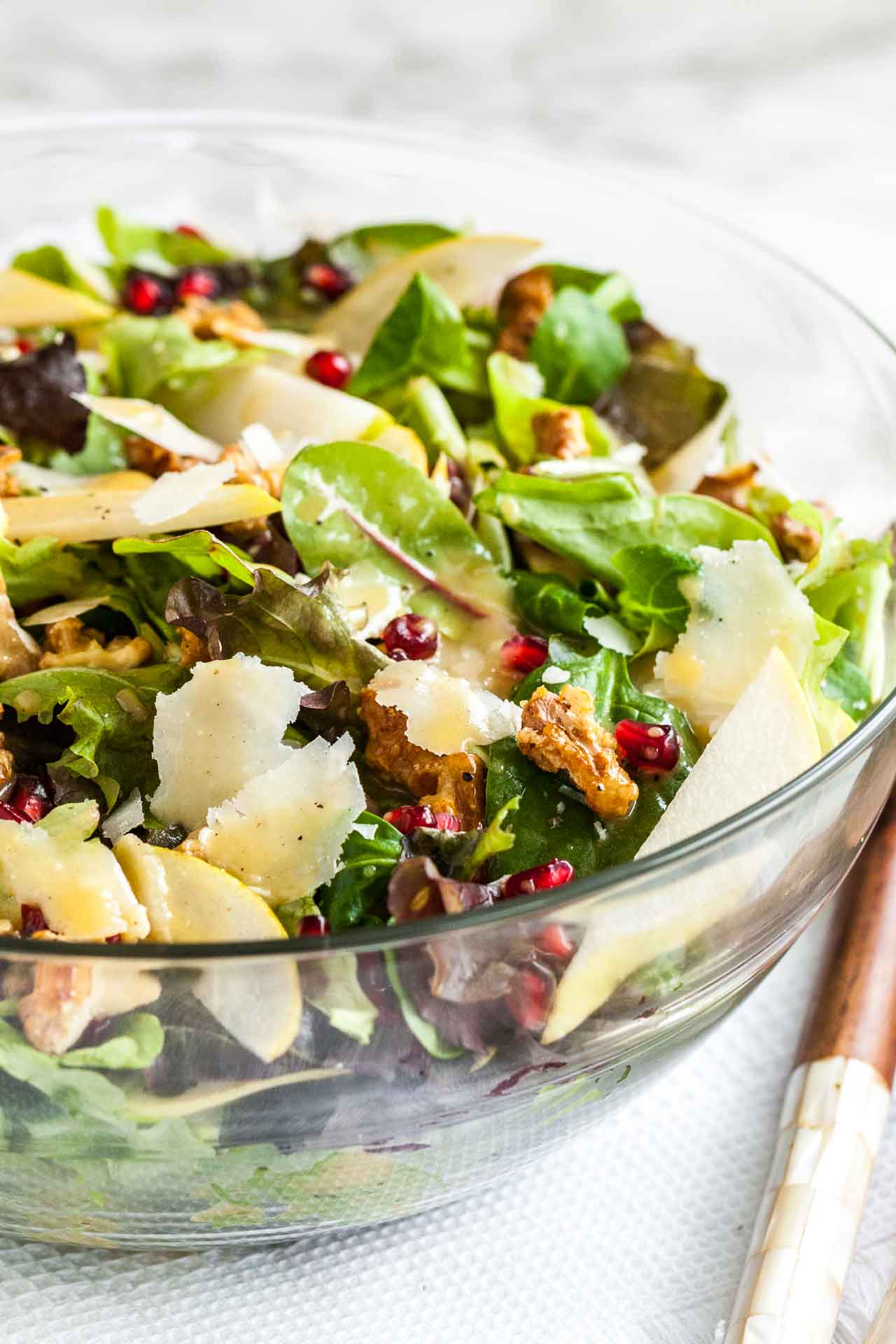 Gros plan d'un bol en verre avec une salade de poire et de grenade avec des noix sur un torchon blanc avec des ustensiles de salade en bois.