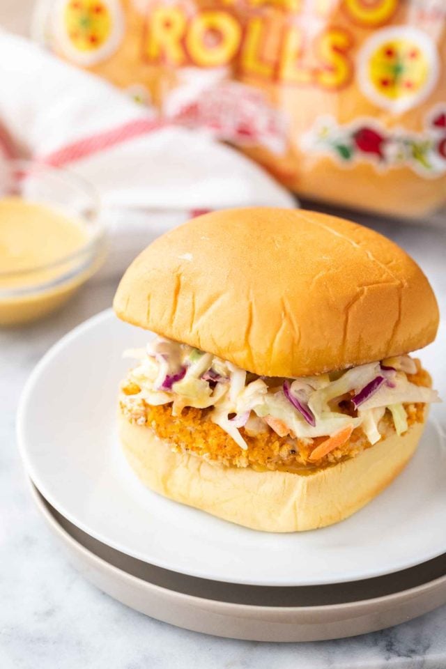 Un burger avec un pavé de poulet frit pané et de la salade de chou sur une assiette blanche à côté d'un bol en verre avec une sauce, une serviette blanche et rouge et un sac de pains à hamburger