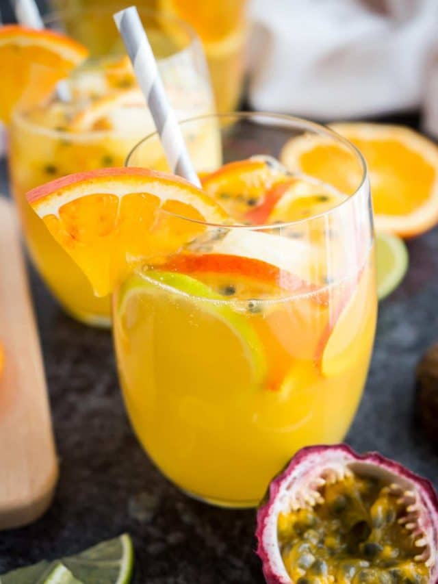 Gros plan d'un verre de sangria blanche tropicale avec des morceaux d'orange, des graines de fruit de la passion et une paille sur une surface sombre. Il y a des tranches d'orange, un autre verre et des fruits de la passion à côté.