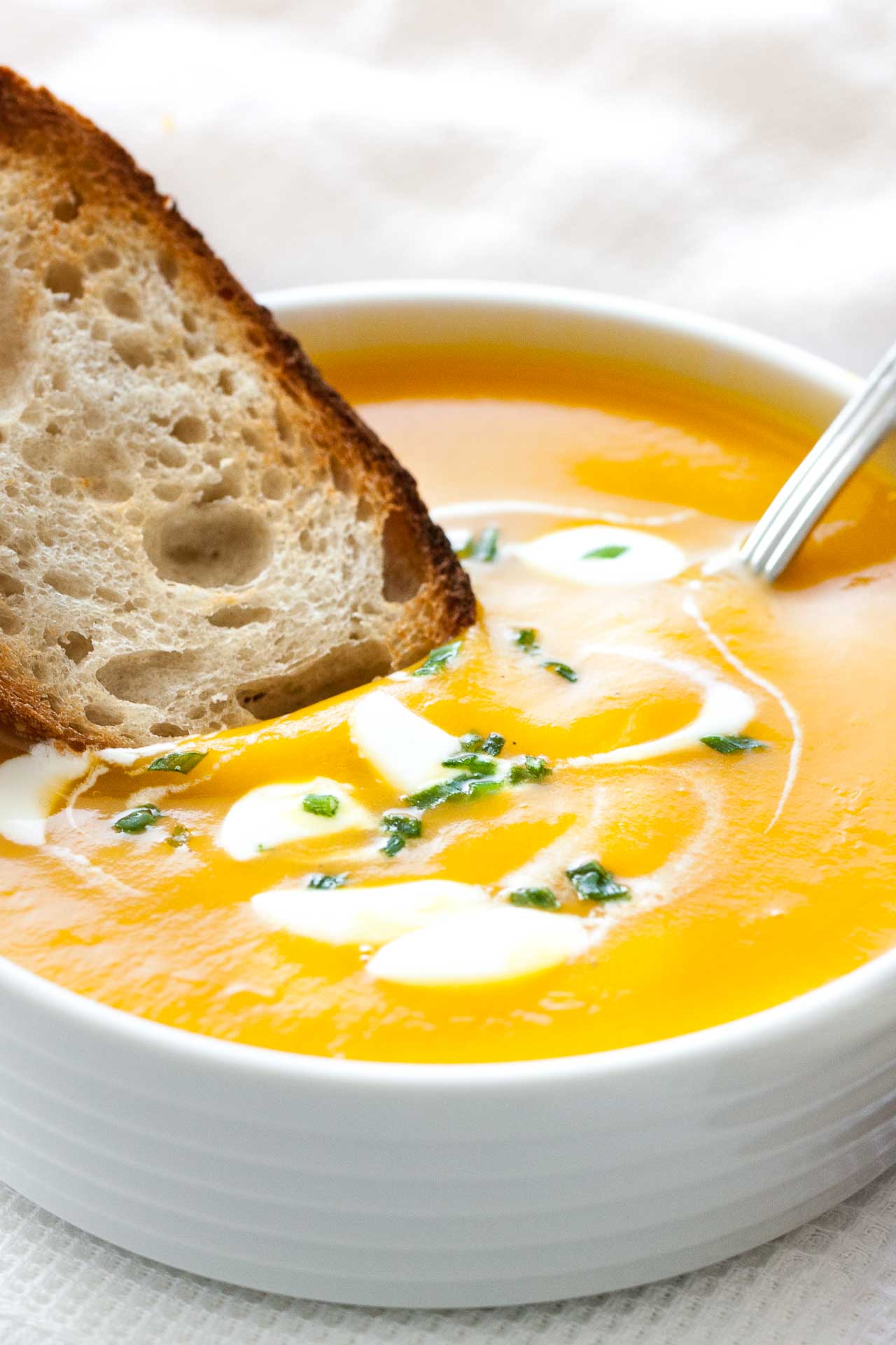 Gros plan d'un bol blanc avec de la soupe de potiron butternut, avec des cuillerées de crème aigre et de la ciboulette, une cuillère dedans et une tranche de pain grillé. Il est posé sur un torchon blanc à côté de quelques tranches de pain grillé.