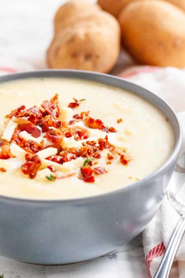 Un bol gris de soupe de pommes de terre garni de miettes de bacon, de bandes de fromage et de thym. Des pommes de terre en arrière-plan.