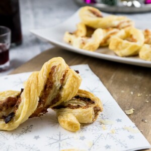 Une serviette festive avec des torsades de pâte feuilletée au bacon et fromage sur une planche à découper en bois. Il y a une assiette rectangulaire blanche en arrière-plan avec d'autres torsades de pâte.