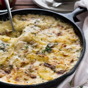 Une poêle en fonte avec des pommes de terre au gorgonzola au gratin et une cuillère qui la sert sur une table en bois. Il y a une serviette blanche à côté et une pile d'assiettes avec des fourchettes en arrière-plan.