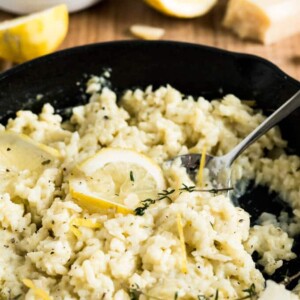 Une poêle en fonte avec du risotto au citron garni de thym et de tranches de citron sur une planche à découper en bois. Il y a des citrons coupés en deux et un morceau de Parmiggiano en arrière-plan.