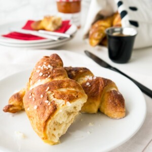 Une assiette blanche avec des croissants bretzel sur une nappe blanche. Il y a plus de croissants, une pile d'assiettes avec des serviettes rouges et des couteaux, une tasse noire d'espresso et un couteau noir en arrière-plan.