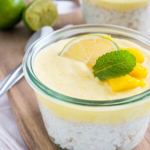 Un pot de pudding de riz à la mangue pour le petit-déjeuner, végan et à la noix de coco, orné d'une tranche de lime, de menthe et de mangue sur une planche à découper en bois. Il y a un autre pot de pudding à l'arrière-plan à côté de quelques cuillères et de limes coupées en deux.