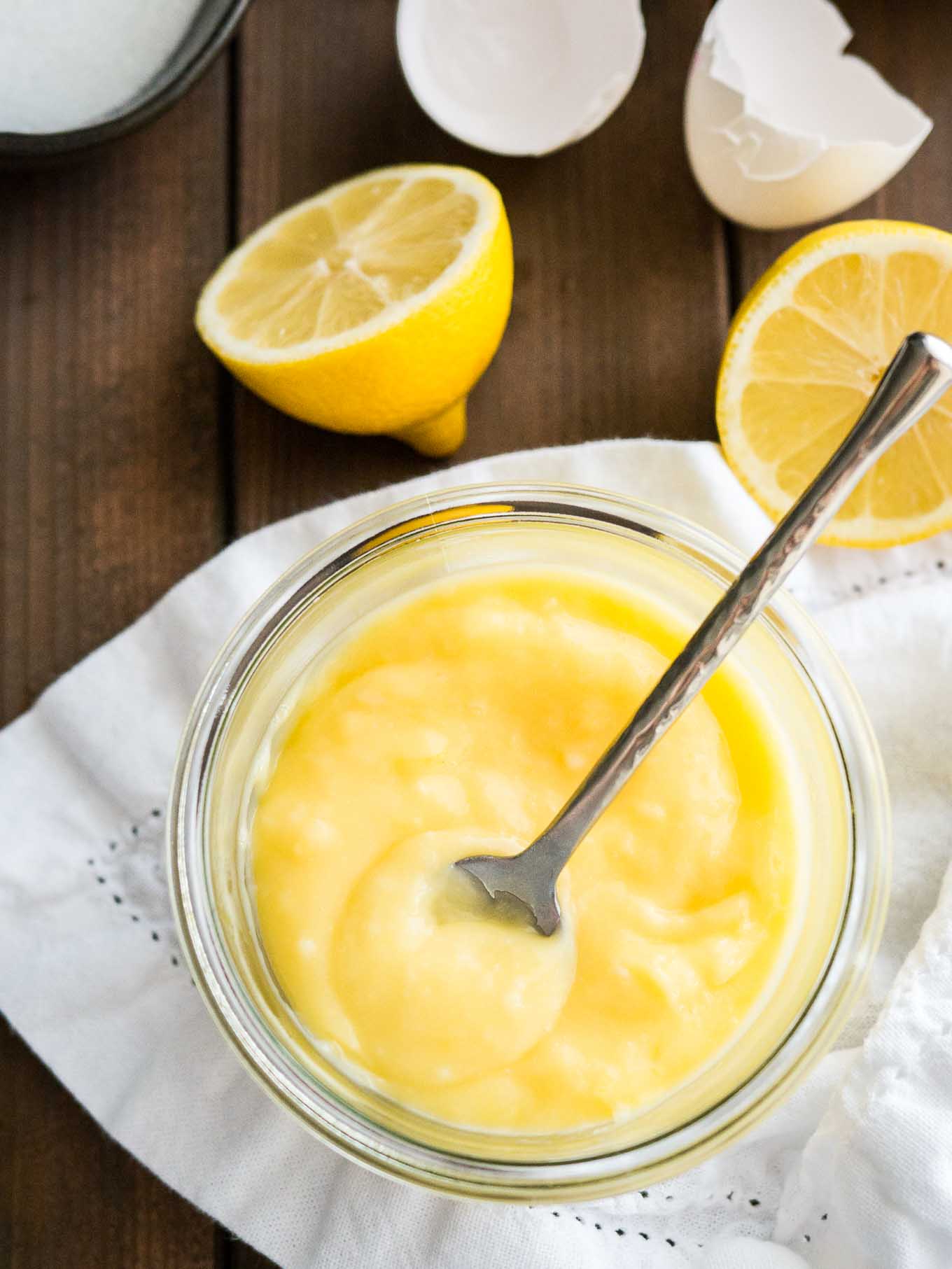 Vue de dessus d'un pot en verre de lemon curd avec une cuillère dedans sur un torchon blanc. Il y a des citrons coupés en deux et des coquilles d'œufs à côté.
