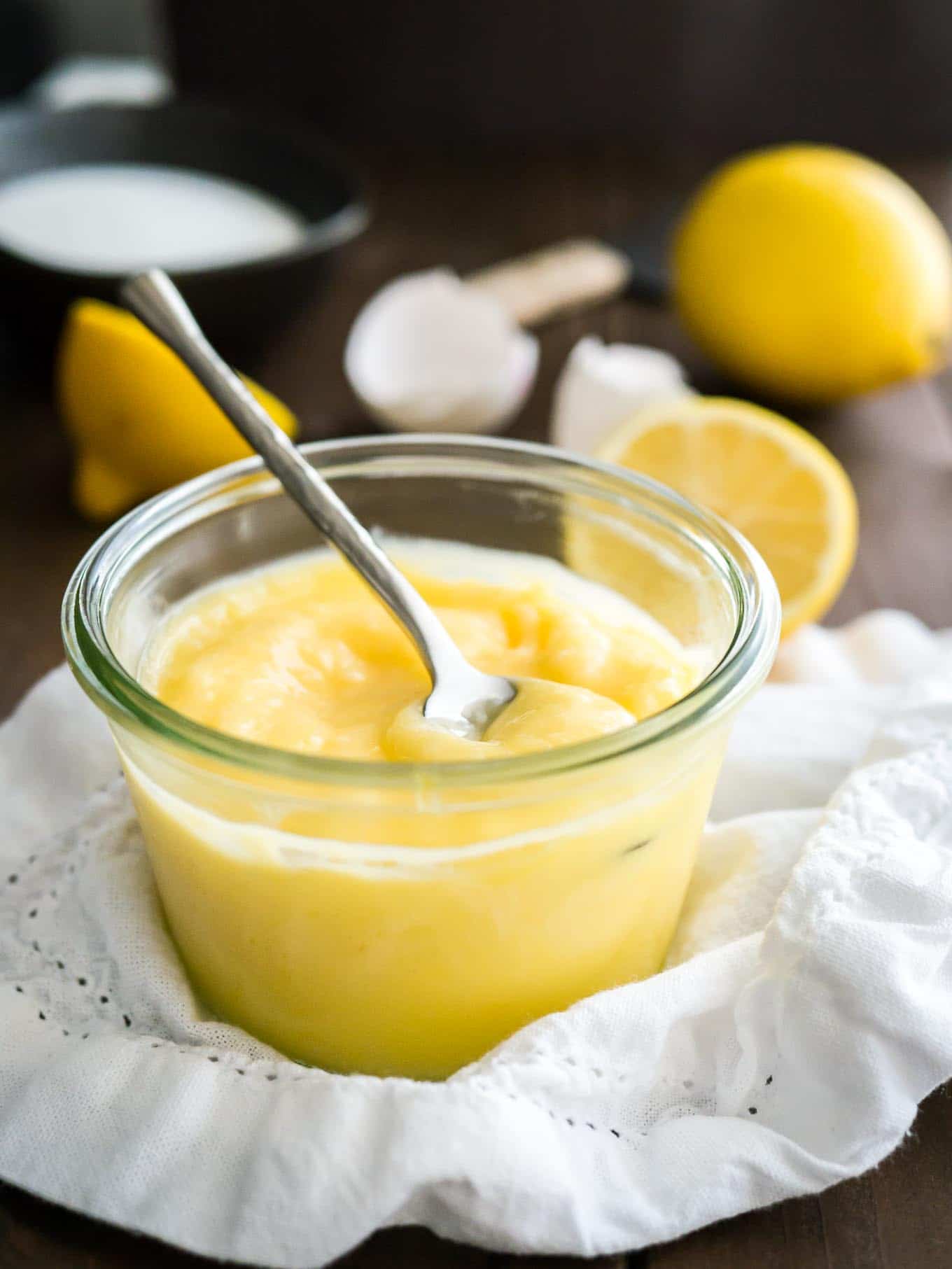 Un pot en verre de lemon curd avec une cuillère dedans sur un torchon blanc. Il y a des citrons coupés en deux et des coquilles d'œufs en arrière-plan.