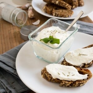 Une assiette blanche avec du pain et une trempette au fromage feta, et un bol en verre avec une trempette au fromage feta et une cuillère dessus sur un torchon gris. Il y a une assiette avec du pain, quelques gousses d'ail et un contenant d'épices en arrière-plan.