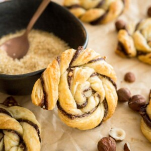 Une danish tordue à la Nutella s'appuyant contre un bol noir avec du sucre brun et une cuillère en bronze garnie de noisettes sur du papier parchemin brun. Il y a plus de danishes autour.