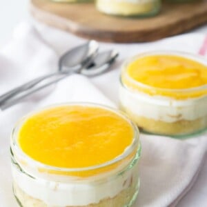 Deux pots avec un dessert aux pêches en conserve sur un torchon blanc avec des cuillères. Il y a une planche à découper avec plus de desserts en arrière-plan.