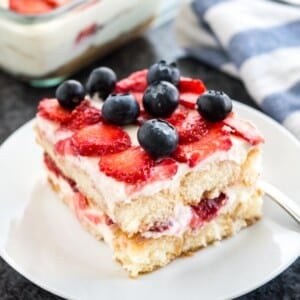 Une assiette blanche avec un morceau de tiramisu aux fraises surmonté de myrtilles. Le reste du tiramisu est dans un plat en verre à l'arrière à côté d'un torchon blanc et bleu.