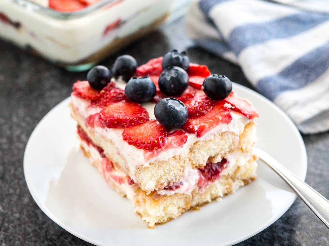 Une assiette blanche avec un morceau de tiramisu aux fraises surmonté de myrtilles. Le reste du tiramisu est dans un plat en verre à l'arrière à côté d'un torchon blanc et bleu.