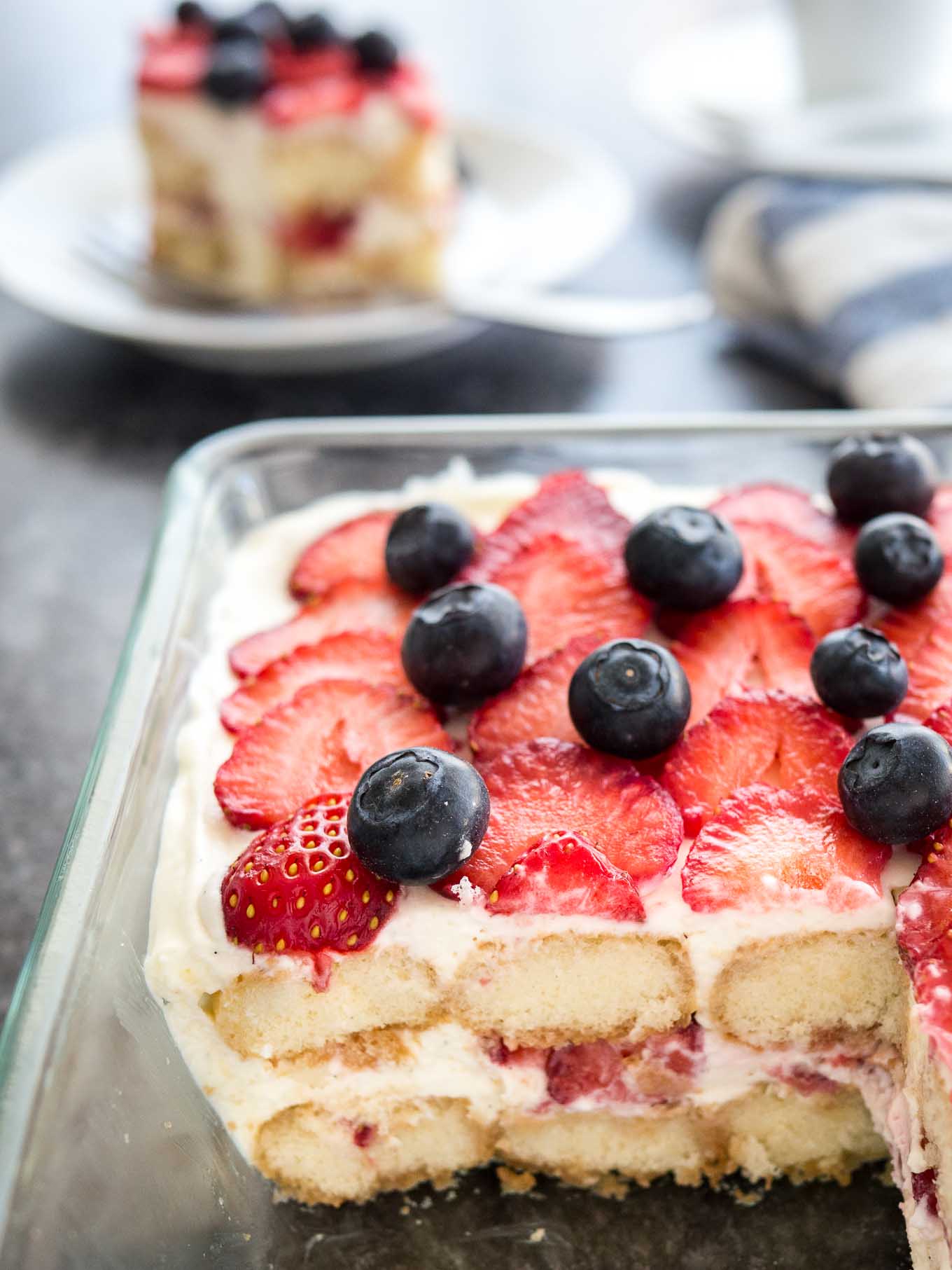 Tiramisu aux fraises dans un plat en verre surmonté de myrtilles. Un morceau du tiramisu a été découpé et se trouve sur une assiette blanche à l'arrière, à côté d'une tasse de café blanche et d'un torchon blanc et bleu.