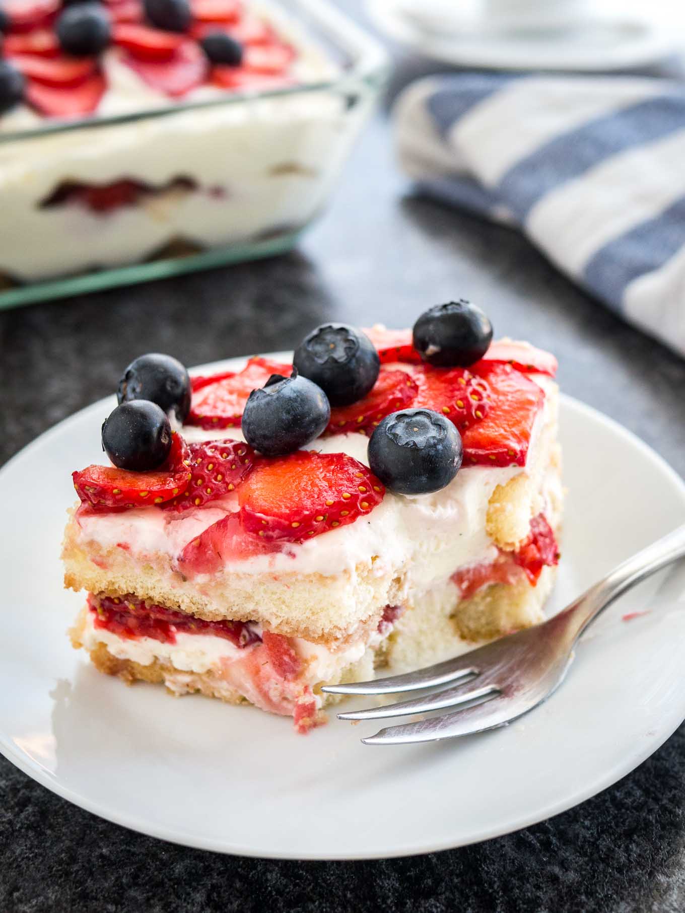 Une assiette blanche avec un morceau de tiramisu aux fraises surmonté de myrtilles. Le reste du tiramisu est dans un plat en verre à l'arrière à côté d'un torchon blanc et bleu. Un morceau du tiramisu a été retiré et une fourchette est posée devant.