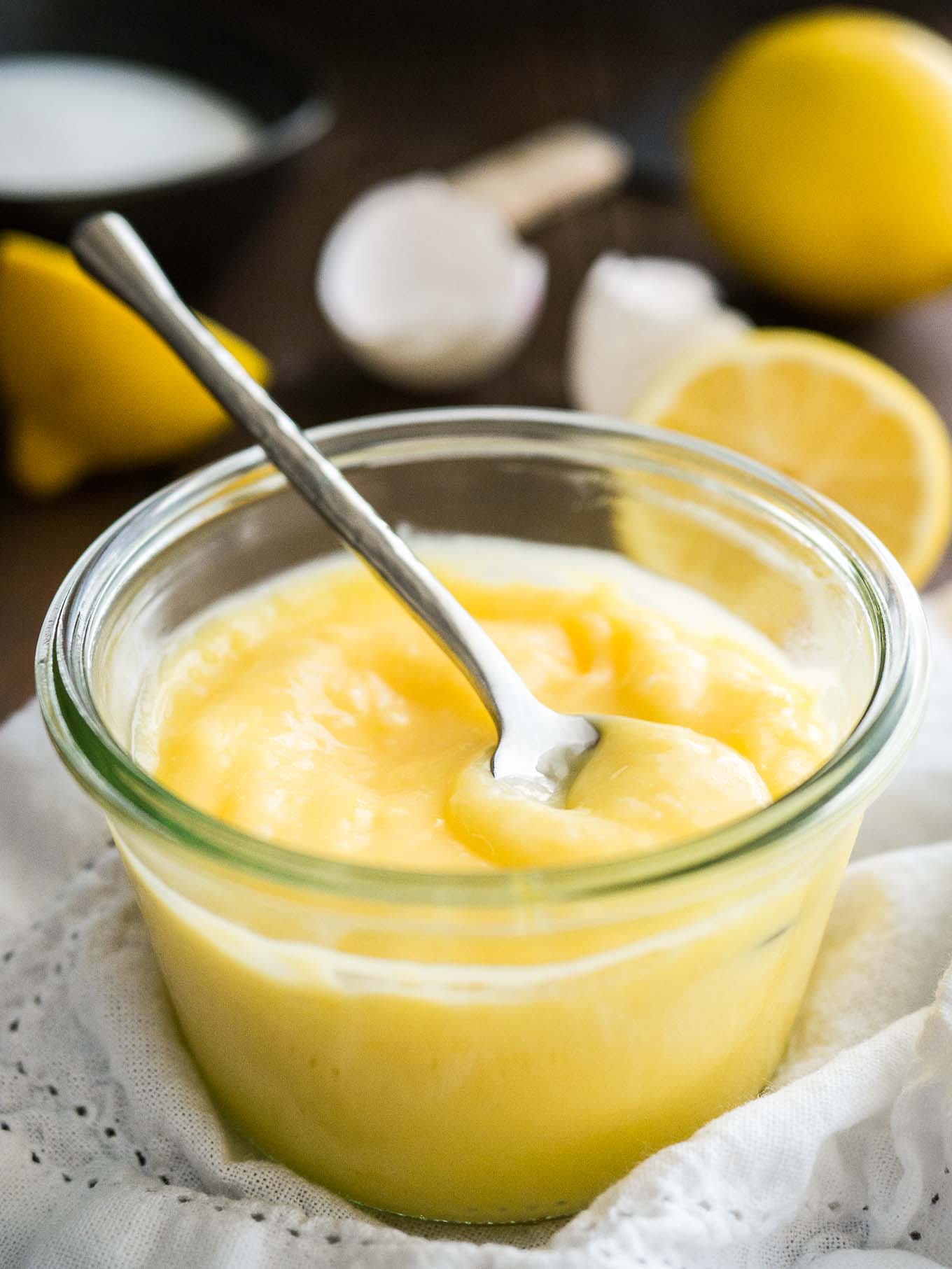 Gros plan sur un pot en verre de lemon curd avec une cuillère dedans sur un torchon blanc. Il y a des citrons coupés en deux et des coquilles d'œufs en arrière-plan.