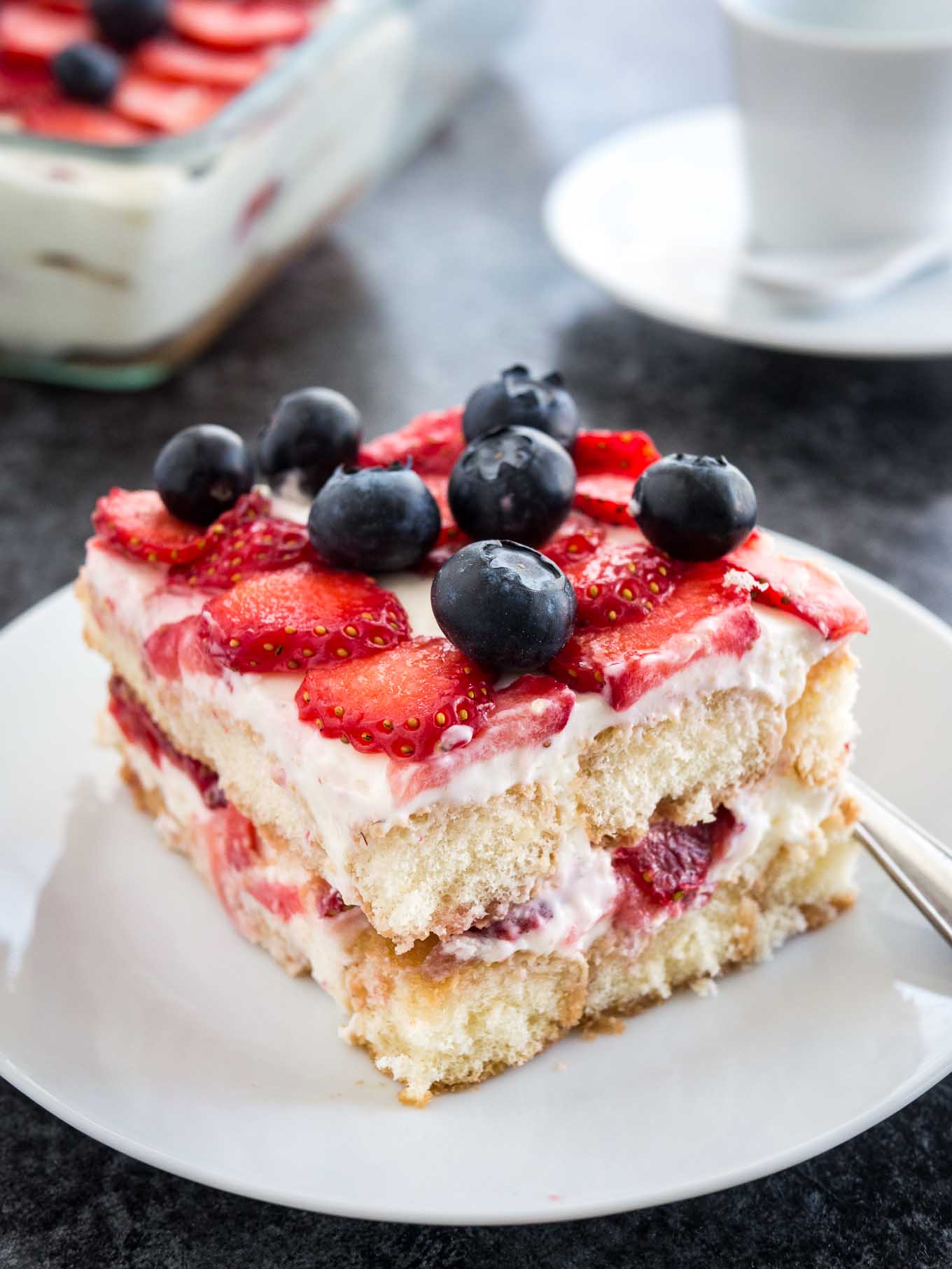 Une assiette blanche avec un morceau de tiramisu aux fraises surmonté de myrtilles. Le reste du tiramisu est dans un plat en verre à l'arrière, à côté d'une tasse de café blanche.